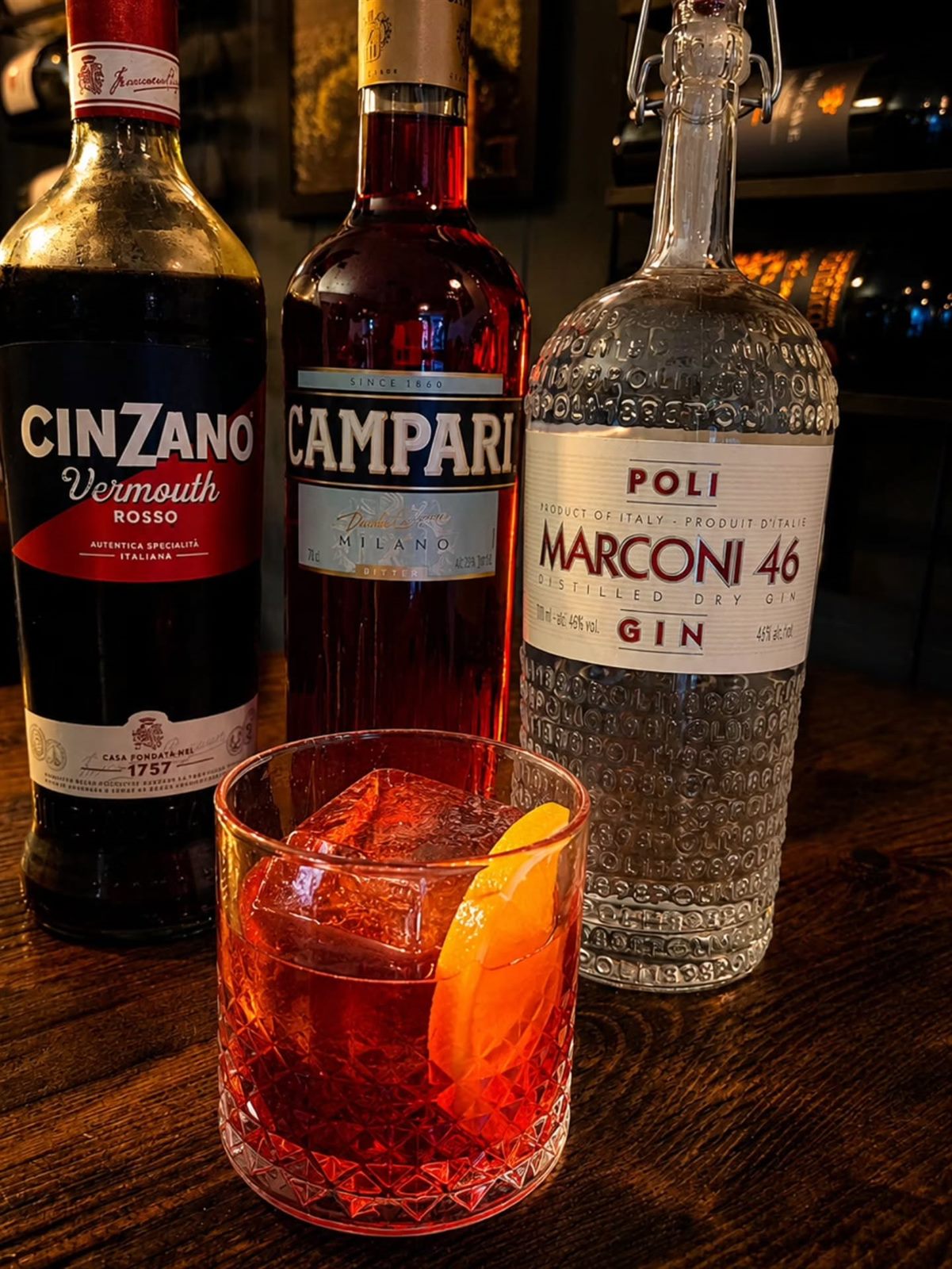 A negroni over ice with an orange peel, beside bottles of Cinzano Rosso vermouth, Campari and Poli Marconi 46 gin