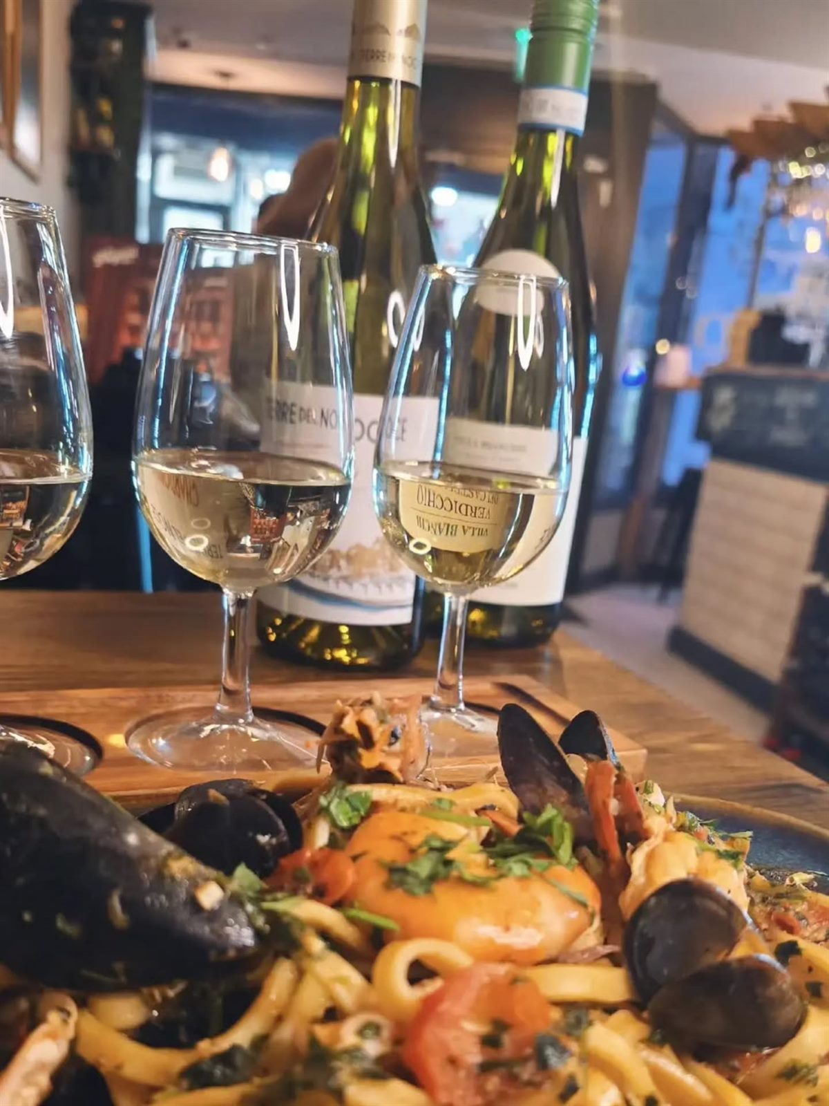 A seafood pasta on the table with three glasses of Italian white wine and their bottles behind
